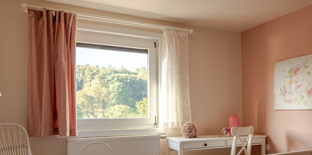 Une pièce avec une fenêtre, un radiateur, un bureau, une chaise et un mur orné d’un tableau représentant des motifs floraux.