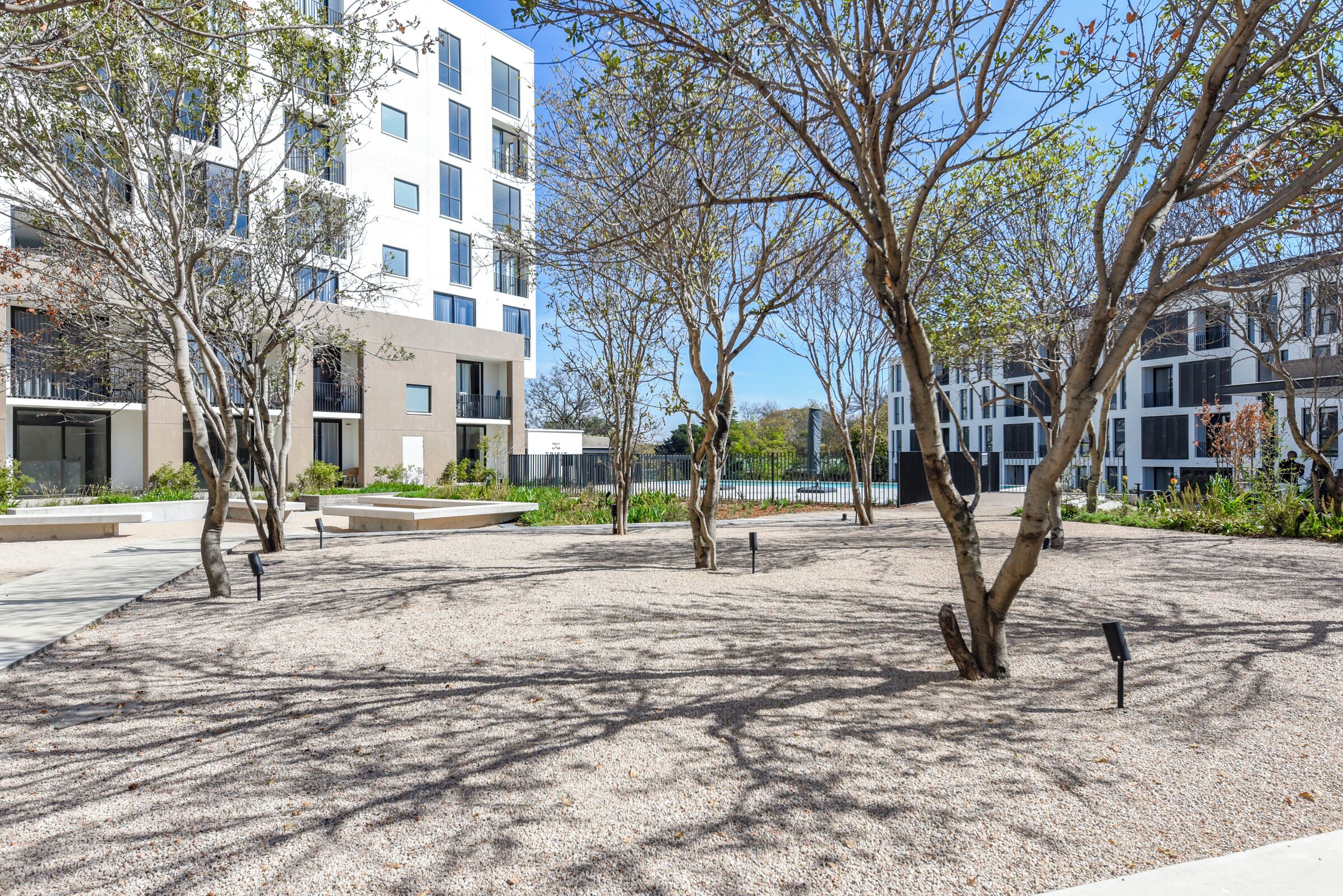 A modern building with multiple floors, surrounded by a landscaped area with trees and gravel paths.
