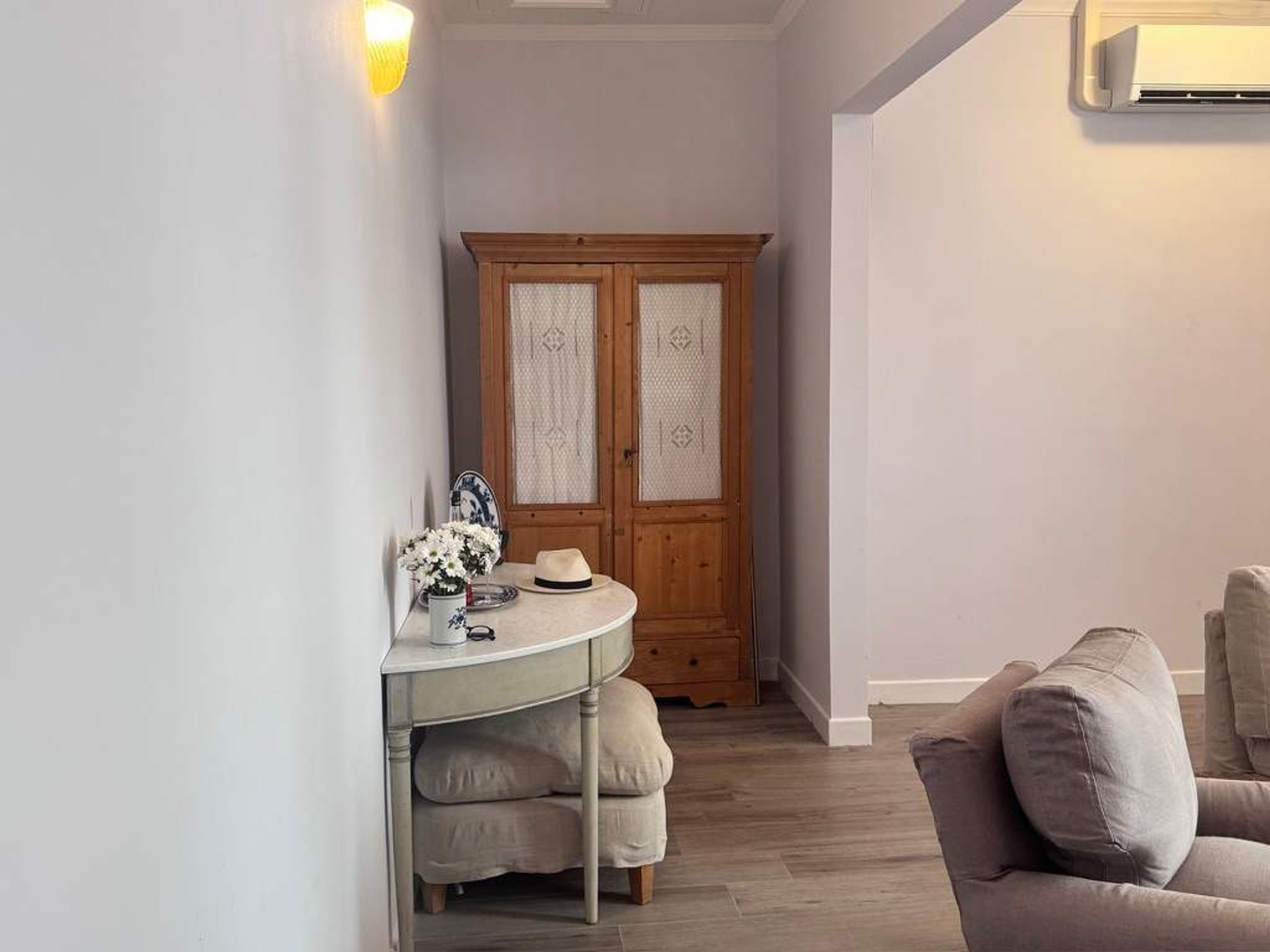 Una habitación con un armario de madera, una mesa redonda con un sombrero y flores, y un sofá gris.