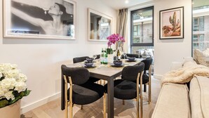 A dining area with a table set for four, surrounded by chairs. There are framed artworks on the walls, a window with a view, and a sofa with a fluffy blanket. - Modern Luxury Apartment in London’s Historic Heart (London)