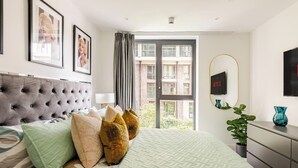 A modern hotel room with a large bed, a grey tufted headboard, multiple pillows, a green quilted bedspread, a large window with curtains, a flat-screen TV mounted on the wall, a potted plant, and a small table with a vase. - Modern Luxury Apartment in London’s Historic Heart (London)