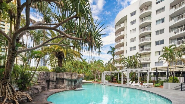 Un área de piscina con borde curvo, rodeada de palmeras y un edificio blanco con balcones.