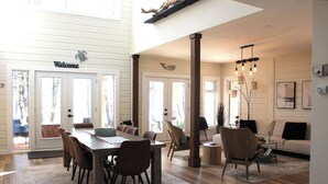 Un amplio salón con una mesa de comedor, sillas, un sofá y una mesa de centro. Hay ventanas grandes que permiten la entrada de luz natural y un cartel con la palabra 'Welcome' sobre las puertas.