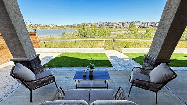 An outdoor patio with a blue table and four wicker chairs, overlooking a body of water and a residential area. - Mahogany Lakefront 2BR Walkout | Trails & Privacy (Calgary)