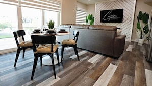 A modern living room with a grey sofa, two black chairs with cushions, a dining table with a plant, and a TV mounted on a wall with a geometric pattern. - Mahogany Lakefront 2BR Walkout | Trails & Privacy (Calgary)