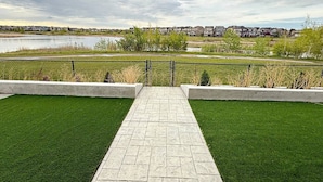A paved pathway leading to a grassy area with a view of a lake and houses in the background. - Mahogany Lakefront 2BR Walkout | Trails & Privacy (Calgary)