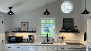 A modern kitchen with white cabinetry, a dishwasher, a stove, and a microwave. There are black pendant lights, a window with a view, and a sign on the wall.