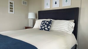 A neatly made bed with a dark blue throw blanket, a white pillow with a geometric pattern, and a plain white pillow. There are framed pictures on the wall above the bed.