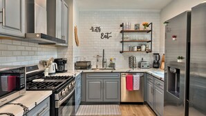 A modern kitchen with grey cabinets, a stainless steel refrigerator, a built-in oven, and a microwave. There is a 'Let's Eat' sign on the wall and a small shelf with decorative items. - Redwood Retreat  Sauna, Plunge, Jacuzzi & Views (Camp Meeker)