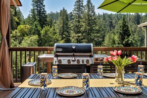 Une terrasse extérieure équipée d’un barbecue Weber, d’une plateforme en bois et d’une table dressée avec des assiettes, des pots et un vase rempli de fleurs.