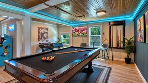 A pool table with a pool cue and balls, a foosball table, and a large TV screen in a room with wooden ceilings and blue LED lighting. - Redwood Retreat  Sauna, Plunge, Jacuzzi & Views (Camp Meeker)