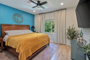 Une chambre avec un lit en bois, un ventilateur de plafond, une fenêtre avec des rideaux et une vue sur la verdure à l’extérieur.