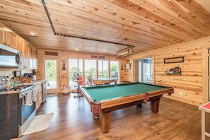 A wooden interior with a pool table, kitchen area, and large windows.