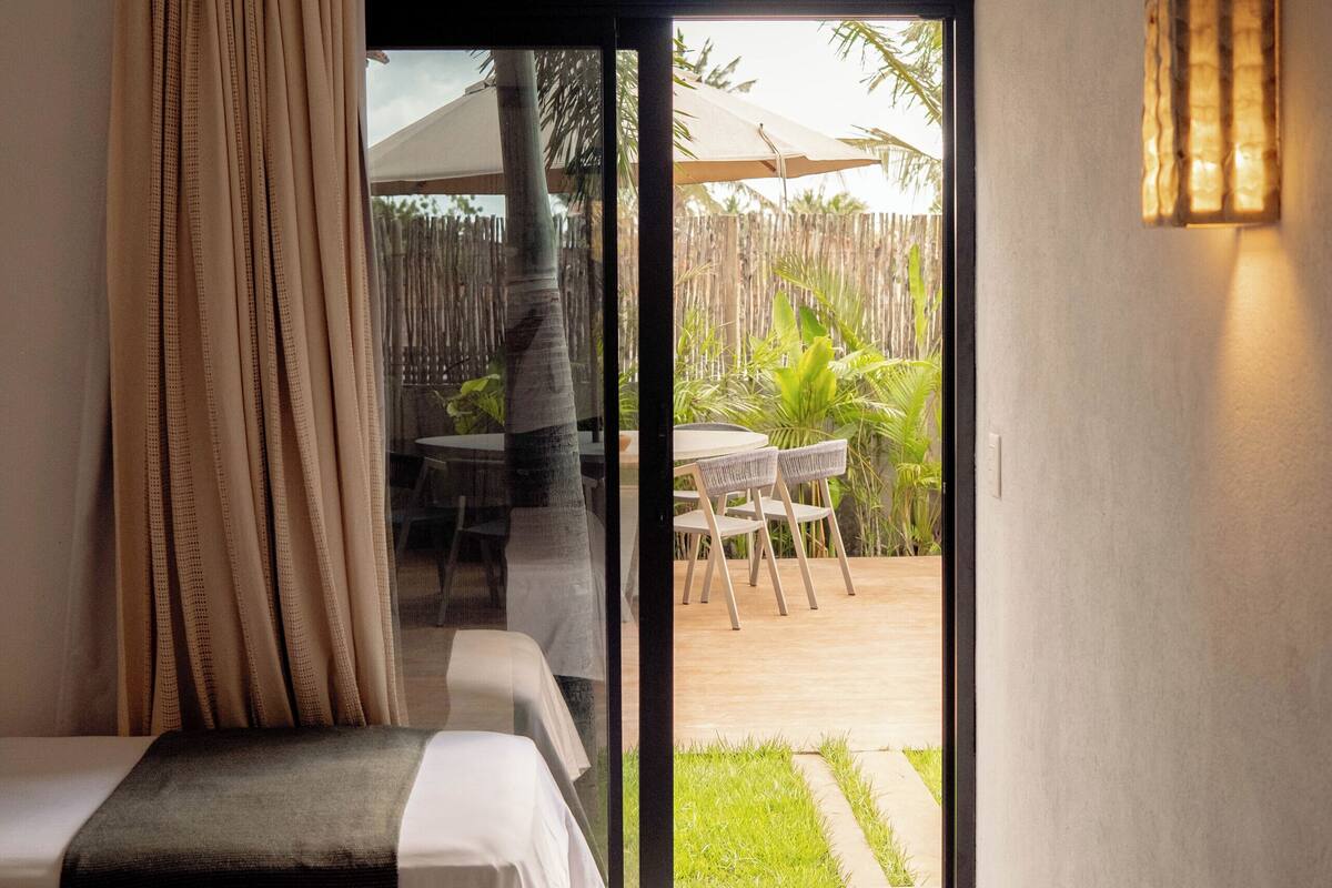 A hotel room with a bed, a window with curtains, a sliding glass door leading to an outdoor area with a table and chairs, and a wall-mounted light fixture.