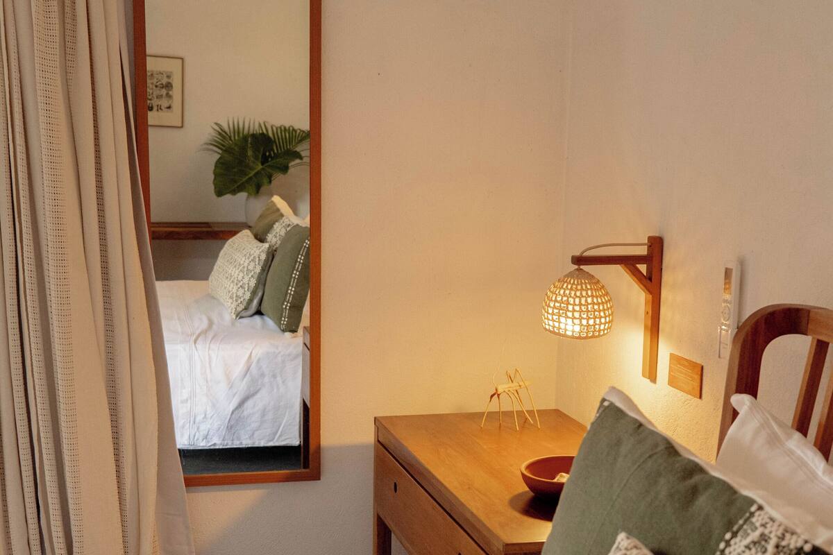 A neatly made bed with white linens, a wooden bedside table with a bowl and a lamp, and a mirror reflecting the room.