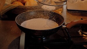 Una cocina con estufa, un colador sobre una sartén y una mesa con pan y una taza.