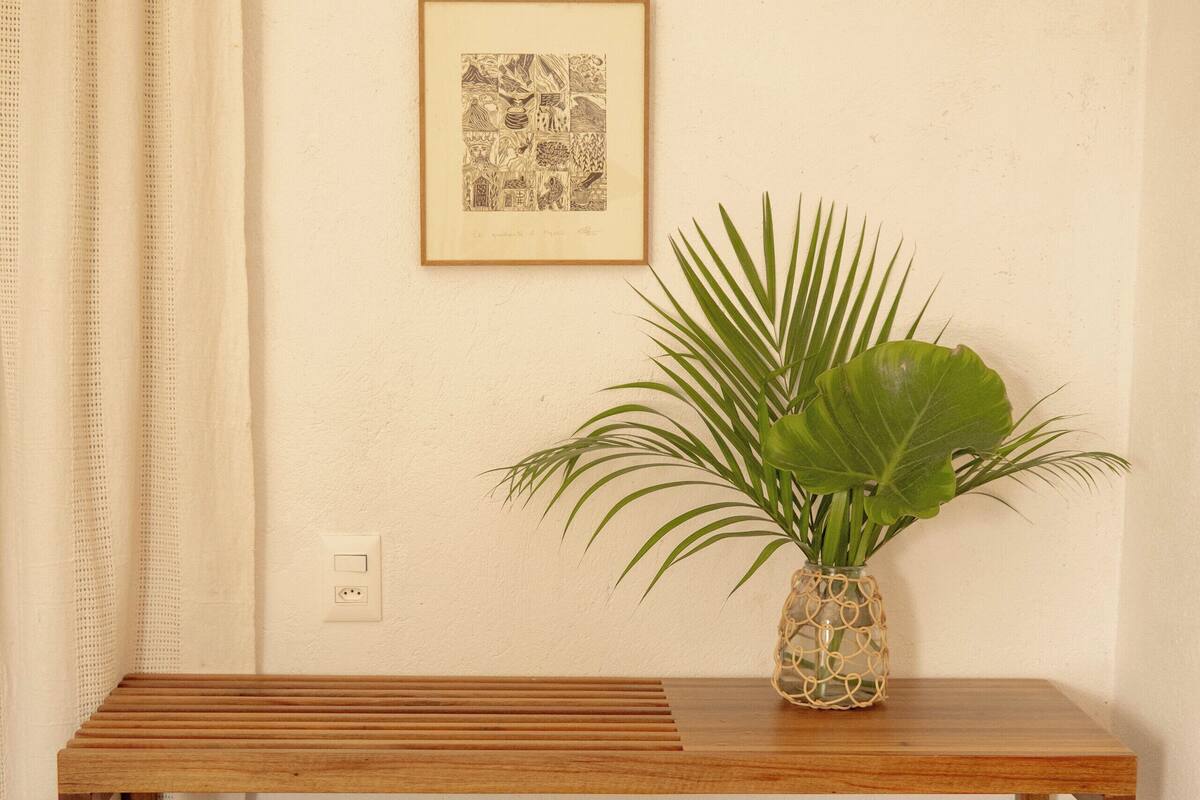 A small wooden desk with a plant in a decorative vase, a framed picture on the wall, and a small stool in front.