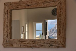 Un miroir rustique, doté d’un cadre en bois, reflète une pièce dans laquelle on aperçoit une porte, une fenêtre, ainsi qu’une vue sur des montagnes et la mer.