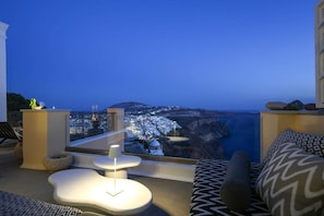 A balcony with a modern design, featuring a white table and a sofa with patterned cushions. A large window offers a view of a coastal town and cliffs.