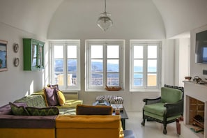 A living room with a high ceiling, large windows with a view of mountains, a green armchair, a yellow sofa, and a fireplace.