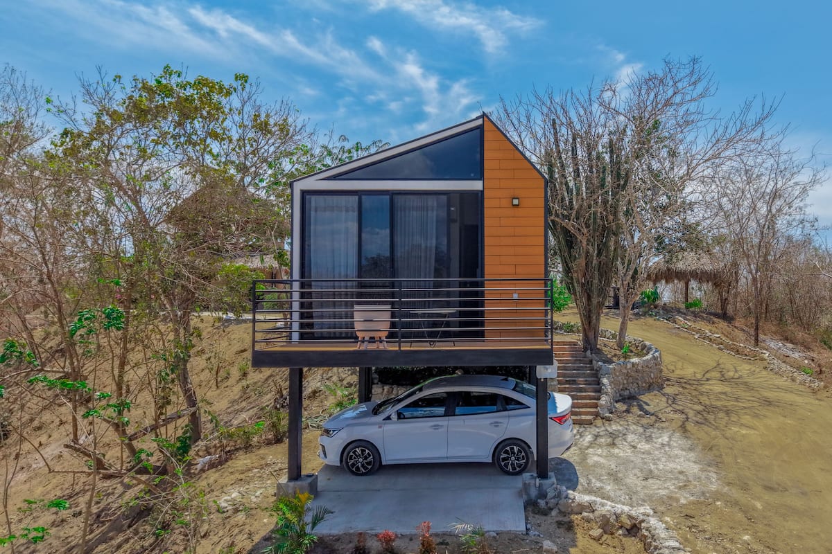 Una casa moderna de dos pisos con un balcón, un coche estacionado al frente y un camino de tierra que lleva a ella.