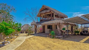 A thatched-roof building with a balcony, surrounded by trees and a sandy ground.