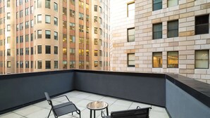 A balcony with two black chairs and a small round table, overlooking a modern building with a mix of brick and glass facade.