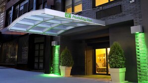 Entrada de un hotel Holiday Inn con toldo de vidrio, fachada de ladrillo y plantas en macetas.