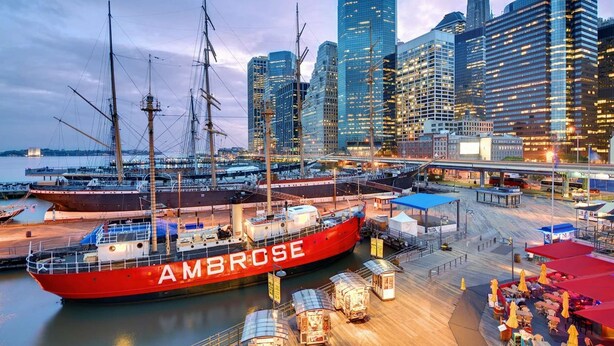 Un gran barco rojo llamado AMBROSE atracado en un puerto costero con edificios modernos al fondo.
