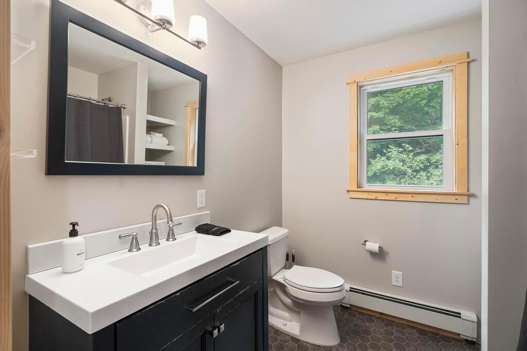 A bathroom with a white sink, dark cabinets, a toilet, and a window with a view of greenery.