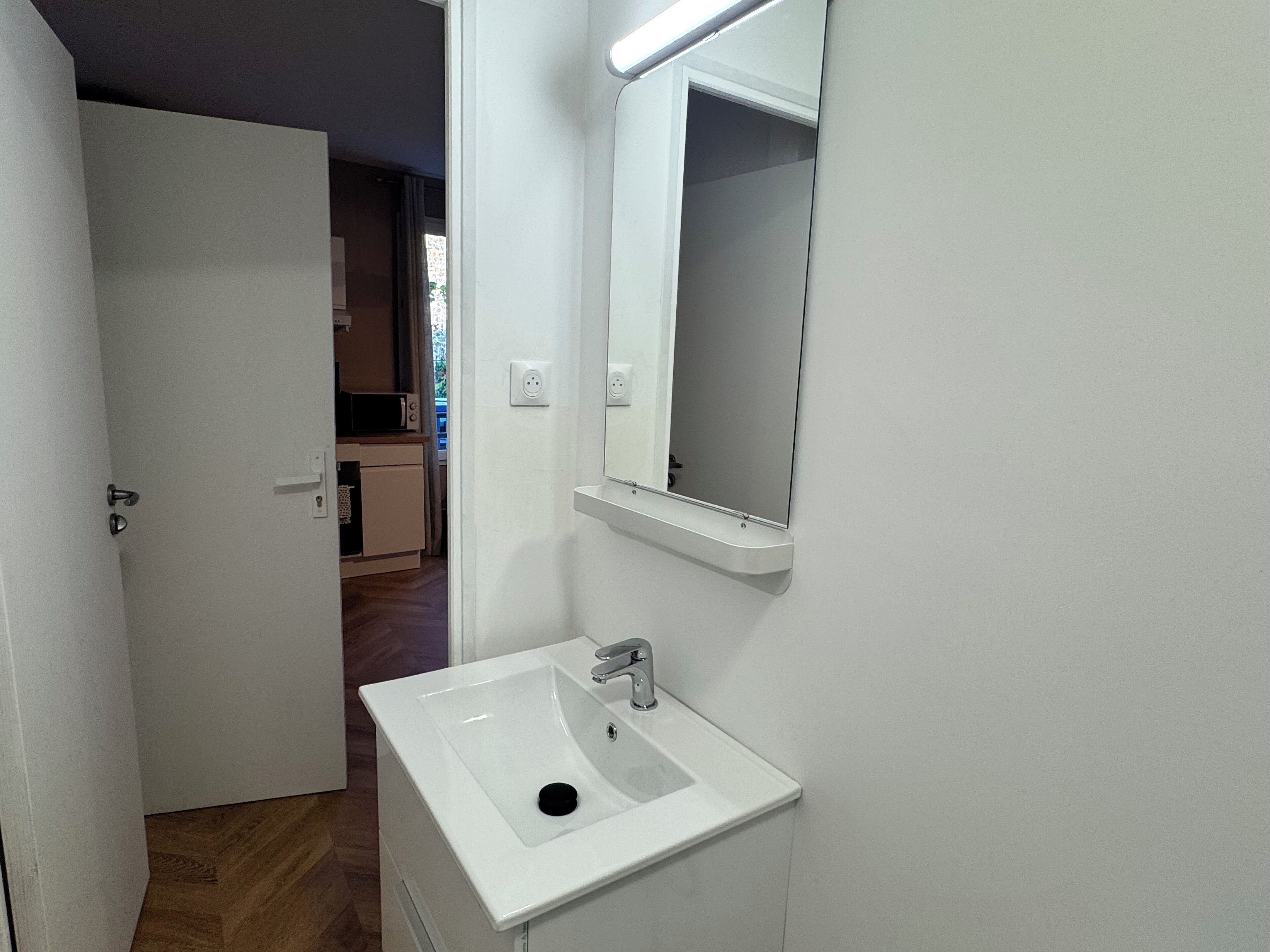 Een smalle badkamer met een wit wastafel, een spiegel en een deur naar een kitchenette.