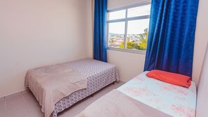 Ein kleines Schlafzimmer mit einem Einzelbett, einem Fenster mit blauen Vorhängen und Blick auf eine Stadtansicht.