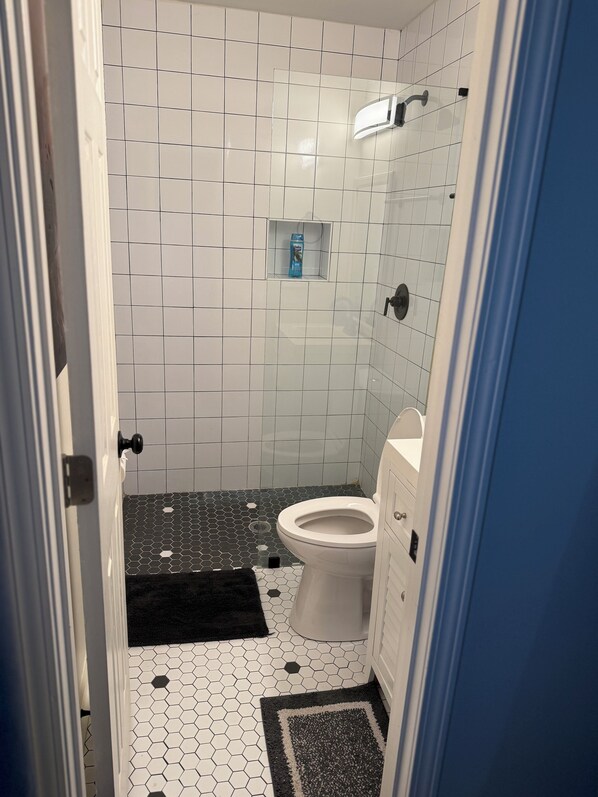 Un baño con paredes de azulejos blancos, un inodoro blanco, un felpudo blanco y negro y un dispensador de jabón azul en un estante pequeño.