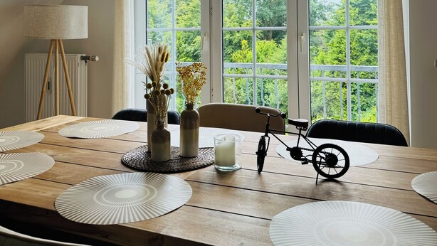 Un coin repas avec une table en bois, des sets de table blancs, une bougie, un vélo et une vue sur la verdure grâce à de grandes fenêtres.