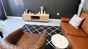 Una sala de estar moderna con un sillón de cuero marrón, una mesa de centro redonda y un sofá naranja. Un televisor de pantalla plana está montado en la pared, y una consola de madera con adornos está colocada contra la pared.