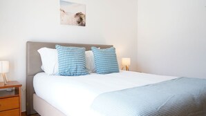 Una cama bien hecha con un juego de almohadas blanco y azul a cuadros, un cabecero gris y una manta de color azul claro. Hay dos mesitas de noche con lámparas, y un cuadro enmarcado con una escena de playa en la pared.