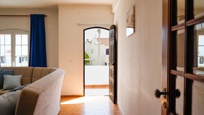 Un pasillo con piso de madera, un sofá beige con cojines, una cortina azul y una puerta que da a un espacio exterior con vistas a edificios y un árbol.