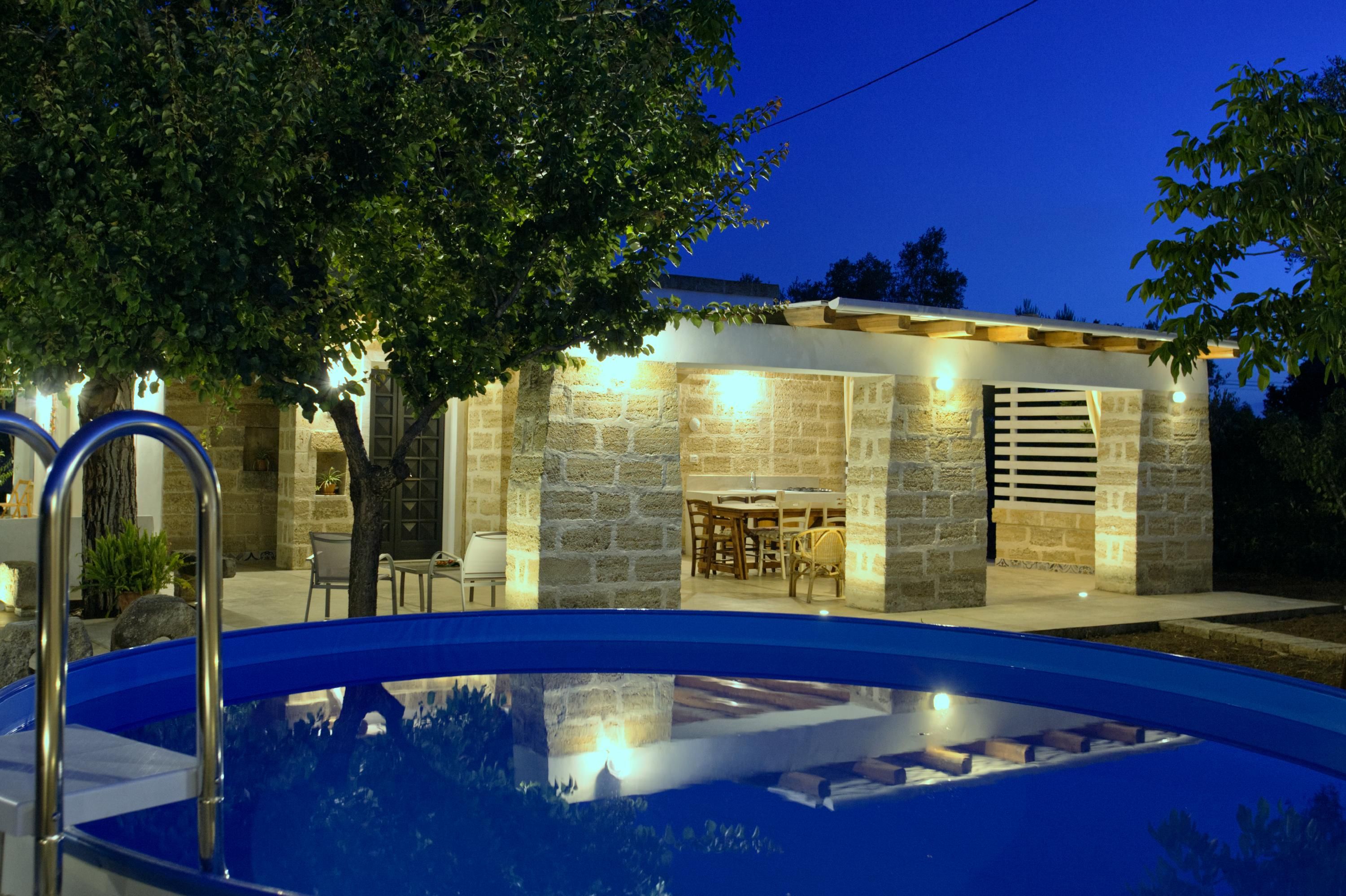 A swimming pool with a stone patio area, a wooden deck, and a stone building with a dining table and chairs.