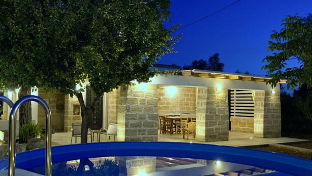 Una piscina con zona de patio de piedra, una terraza de madera y un edificio de piedra con una mesa y sillas.