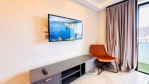 A modern hotel room with a flat-screen TV mounted on the wall, a grey entertainment unit, a brown leather chair, and a large window with curtains. - Newly renovated, charming studio in central Jeffreys Bay with AC, WiFi, parking (Jeffreys Bay)