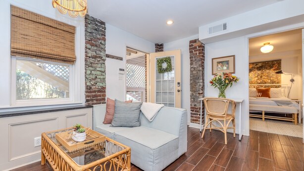 A cozy living room with a light blue sofa, a wicker coffee table, and a wicker chair. There is a brick wall, a wooden door, and a window with a white blind.