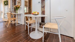 A modern kitchen with white cabinetry, a round dining table, and two chairs. There is a fruit bowl on the table and a vase with flowers on the wall.
