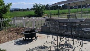Eine Terrasse im Freien mit rundem Tisch und Stühlen, einer Feuerstelle und einem weißen Zaun aus Holzlatten.