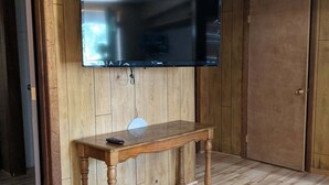 Ein Zimmer mit Holzboden, einem an der Wand befestigten Fernseher, einem Holztisch und einer Tür.