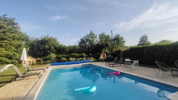 A rectangular swimming pool with clear blue water, surrounded by a wooden deck. There are lounge chairs and a small table with chairs on the deck.