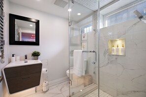 Une salle de bain moderne avec une douche à l’italienne en verre, un meuble-lavabo en bois foncé et un mur de douche recouvert de carreaux de marbre.