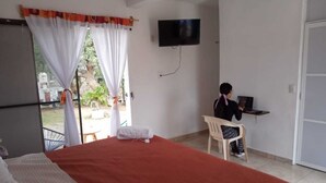 Une chambre à coucher avec un lit, un bureau avec un ordinateur, un téléviseur fixé au mur et une fenêtre avec des rideaux.