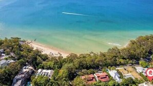 An aerial view of a coastal area with a beach, dense greenery, and several buildings.