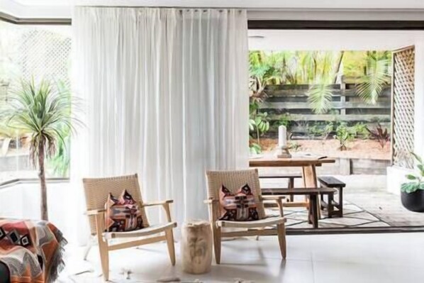 Una habitación con dos sillas de mimbre, una mesa de madera y vistas a un jardín a través de una ventana con cortinas blancas.