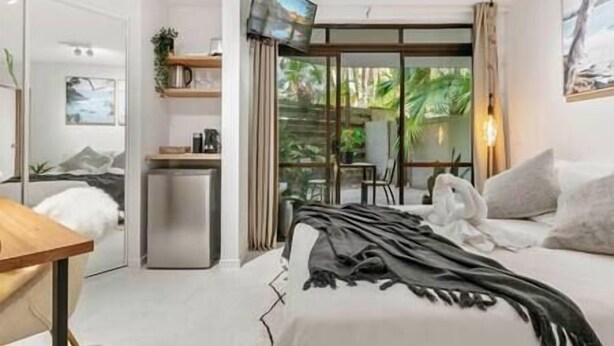 A modern bedroom with a bed, a small desk, a refrigerator, and a sliding glass door leading to a balcony.